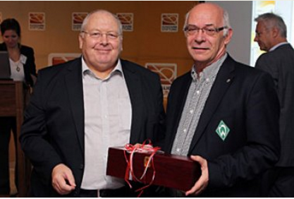 Zwei Männer im Anzug stehen sich gegenüber und halten ein Geschenk mit roter Schleife. Im Hintergrund ist ein Teil eines Konferenzraumes zu sehen.