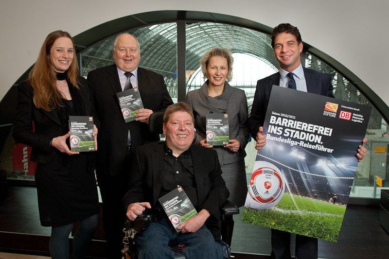 Gruppe von Menschen präsentiert das "Barrierefrei ins Stadion"-Reiseführer-Heft. Vordergrund: Mann in Jeans und Jacke hält den Reiseführer in die Kamera. Hinter ihm stehen vier weitere Personen mit dem gleichen Heft in den Händen, vor einer Glasfassade mit Stadion im Hintergrund. Im Logo sind die DFL Stiftung und die Deutsche Bahn zu sehen.