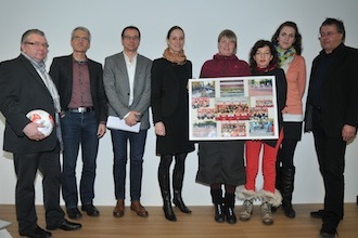 Eine Gruppe von acht Personen steht zusammen und hält ein großes Poster mit Fotos von Fußballaktivitäten hoch. Die Personen sind unterschiedlich gekleidet, von formell bis leger.