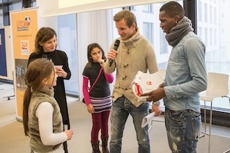 Stefan Aigner und Moussa Quattara stehen auf einer Weihnachtsfeier der DFL Stiftung im Haus des Buches mit einem jungen Mädchen. Aigner hält ein Mikrofon und scheint mit dem Mädchen zu sprechen, während Quattara neben ihm steht. Im Hintergrund sind weitere Personen und Möbel erkennbar.