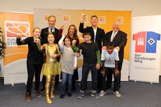 Eine Gruppe von sieben Personen steht lächelnd vor einer hellen Wand mit dem Logo der DFL Stiftung und dem Schriftzug "Der Hamburger Weg - Hamburg beflügelt". Von links nach rechts sind zu sehen: ein Mädchen, eine in Gold gekleidete Person, ein weiteres Mädchen, ein Junge, ein Mann im Anzug, eine Frau und ein Junge im Fußballtrikot. Alle haben die Arme gehoben.