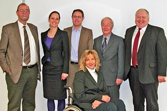 Eine Gruppe von sechs Personen posiert für ein Porträt. Eine Frau im Rollstuhl sitzt in der Mitte, umgeben von fünf weiteren Personen in formeller Kleidung.