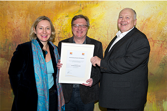 Drei Personen bei der Übergabe einer Urkunde. Links eine Frau mit blonden Haaren und Schal, in der Mitte ein Mann mit Brille, der die Urkunde entgegennimmt, und rechts ein Mann mit Anzug, der die Urkunde überreicht.