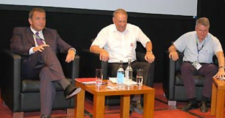 Drei Männer in Sessel sitzen auf einer Bühne, zwischen ihnen steht ein kleiner Tisch mit Wasserflaschen und Gläsern.