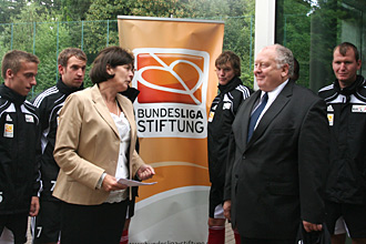 Eine Frau mit Blazer und weiße Bluse spricht mit einem älteren Herrn im Anzug. Im Hintergrund stehen mehrere Fußballspieler in Trainingsanzügen. Im Hintergrund ist ein Banner mit dem Logo der Bundesliga-Stiftung zu sehen.