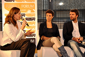 Eine Frau mit Mikrofon auf einem Podium, flankiert von zwei weiteren Personen, während eines Fußball-Forums der DFL Stiftung. Im Hintergrund ein Banner mit der Aufschrift "Fußballforum".