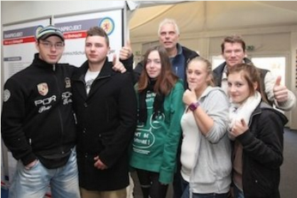 Eine Gruppe junger Menschen steht lächelnd und mit Daumen hoch vor einer Werbetafel im Hintergrund.