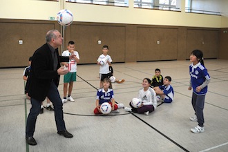 Ein Mann in dunklem Anzug steht in einer Sporthalle und jongliert mit einem Fußball, während ihn eine Gruppe von Kindern umringt. Die Kinder halten ebenfalls Fußball in den Händen und schauen zum Mann auf. Im Hintergrund sind die kahlen Wände der Sporthalle zu sehen. Das Bild zeigt eine Aktion der DFL Stiftung "Fußball trifft Kultur" an der Gemeinschaftsgrundschule an der Leipziger Straße in Gelsenkirchen.