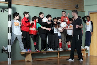 Eine Gruppe von Kindern und Jugendlichen steht in einer Turnhalle auf einer Bank und posiert für ein Foto. Ein Mann steht rechts daneben und lächelt. Im Vordergrund halten einige Kinder Fußballbälle. Es handelt sich um eine Szene des Projekts "Fußball trifft Kultur" der DFL Stiftung in der Albert-Schweitzer-Schule in Frankfurt am Main.