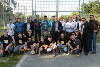 Eine Gruppe von Erwachsenen und Jugendlichen steht und hockt auf einem Sportplatz vor einem Maschendrahtzaun. Im Vordergrund knien und hocken junge Menschen in T-Shirts und Sportkleidung, während im Hintergrund Erwachsene in formellerer Kleidung stehen.