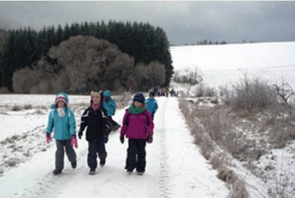 Eine Gruppe von Kindern wandert auf einem verschneiten Feldweg, mit einem dunklen Wald im Hintergrund.