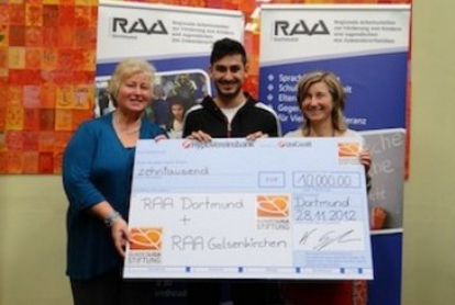Zwei Frauen und ein junger Mann halten einen übergroßen Scheck mit der Aufschrift "40.000 EUR" und den Logos "RAA Dortmund" und "RAA Gelsenkirchen" sowie dem Datum "28.11.2012". Im Hintergrund ein Banner mit dem Logo "RAA".