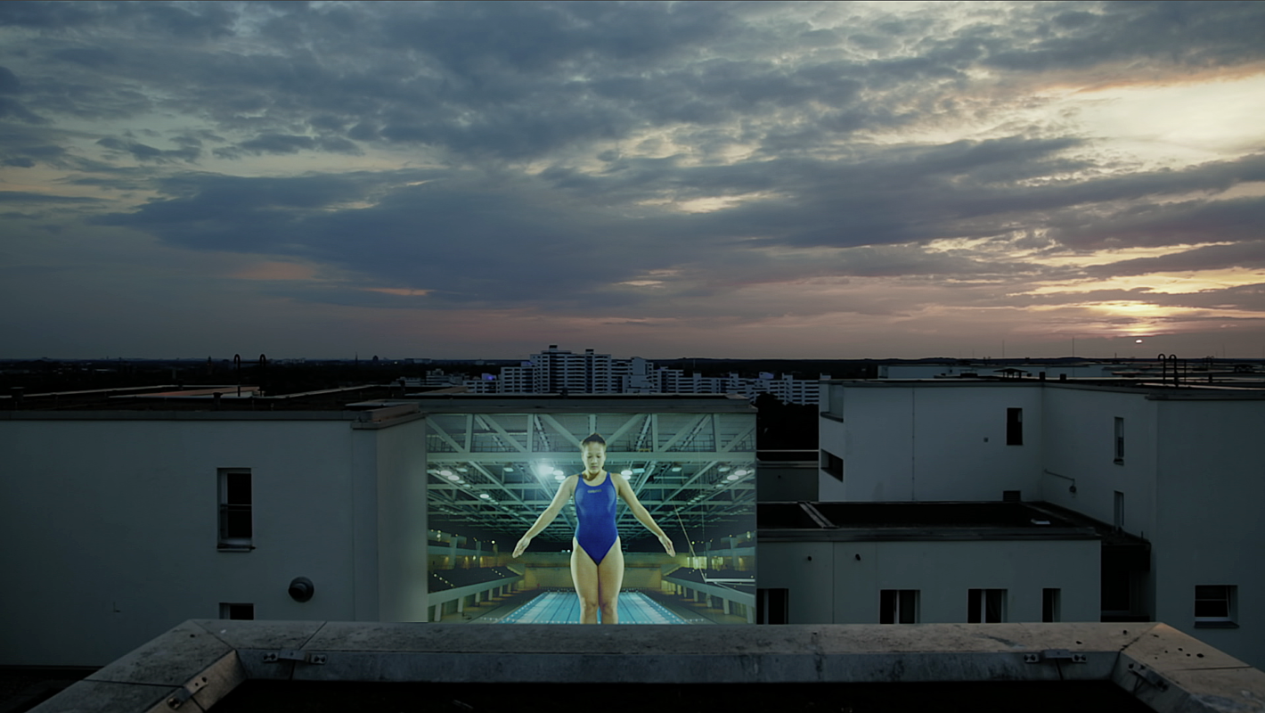 Eine Frau in Sportkleidung steht auf einem riesigen LED-Screen auf dem Dach eines Gebäudes, im Hintergrund eine Skyline bei Sonnenuntergang.