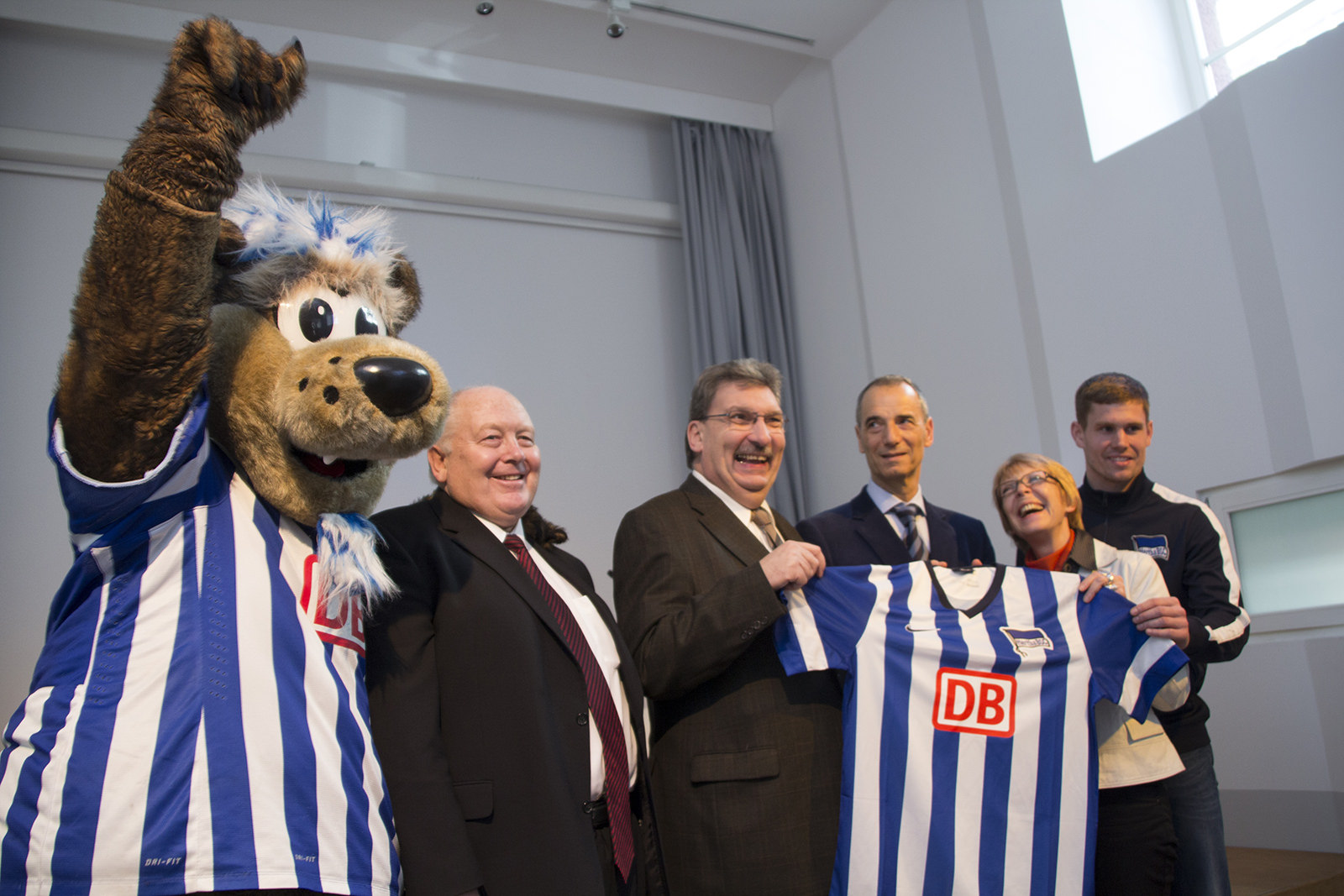 Eine Gruppe von sechs Personen steht lächelnd in einem hellen Raum. Links steht eine Maskottchenfigur eines Bären mit blau-weiß gestreiftem Trikot. Daneben stehen vier Männer und eine Frau im Anzug, die ebenfalls ein blau-weiß gestreiftes Trikot mit dem Logo der Deutschen Bahn präsentieren. Im Hintergrund befindet sich ein großes Fenster.