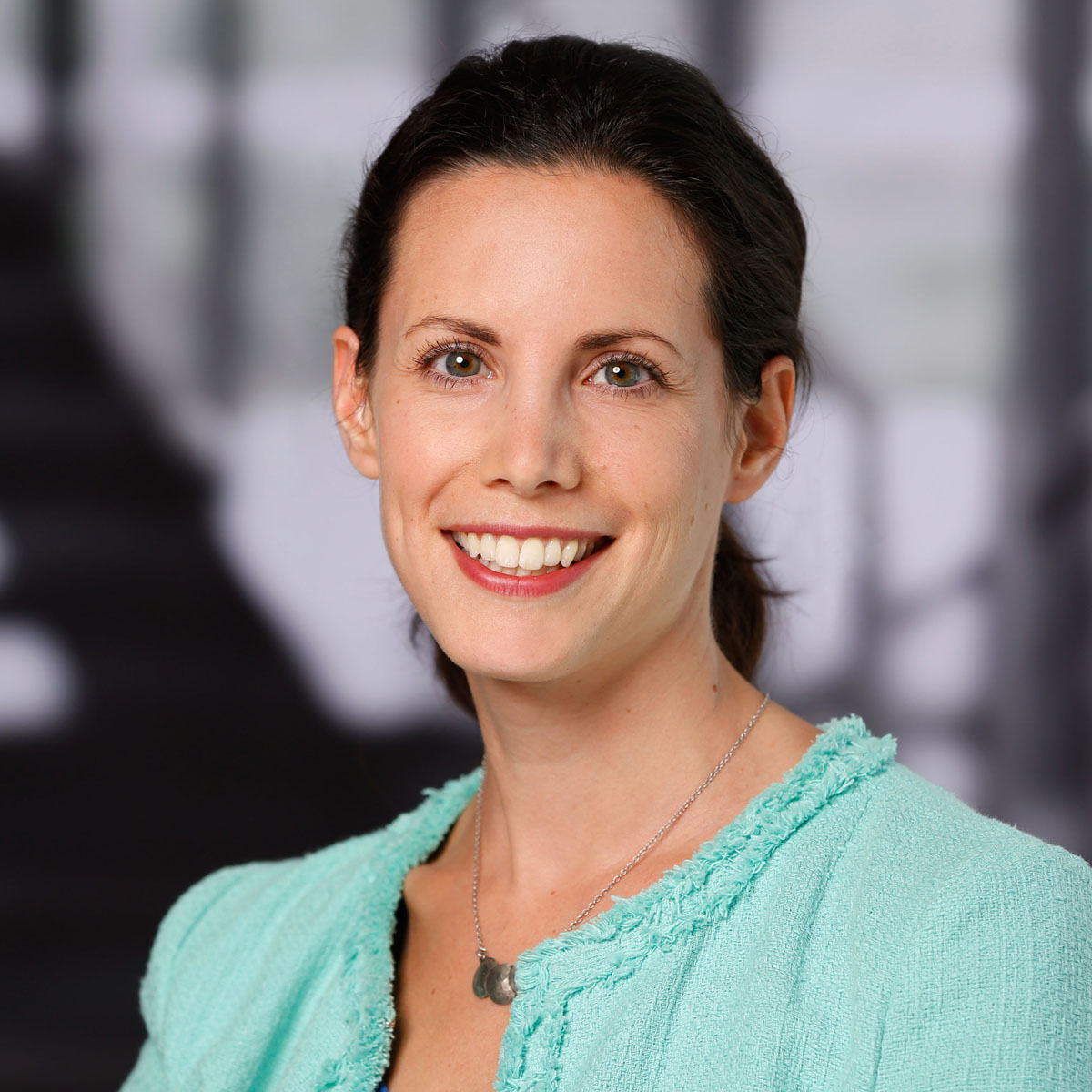 Lachende Frau mit braunen Haaren, trägt ein türkisfarbenes Oberteil und eine feine Kette.