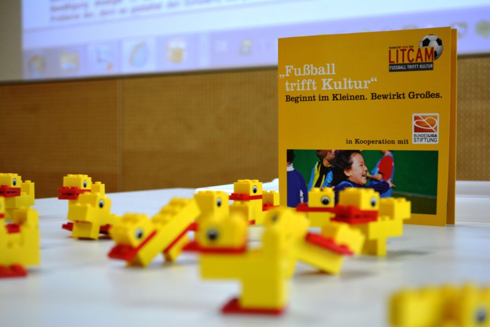 Eine Ansammlung gelber Quietscheentchen steht vor einem aufgestellten Banner mit der Aufschrift "Fußball trifft Kultur - Beginnt im Kleinen. Bewirkt Großes." und dem Logo der Bundesliga-Stiftung.