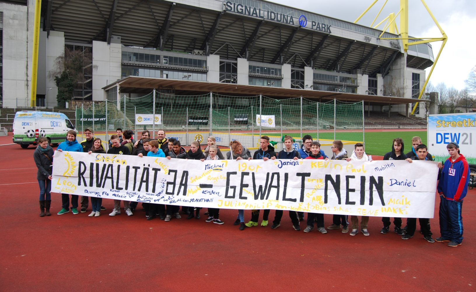 Eine Gruppe von Menschen steht auf einer roten Tartanbahn vor dem Signal Iduna Park und hält ein großes Banner mit der Aufschrift "RIVALITÄT JA – GEWALT NEIN". Im Hintergrund ist das Fußballstadion des BVB zu sehen.
