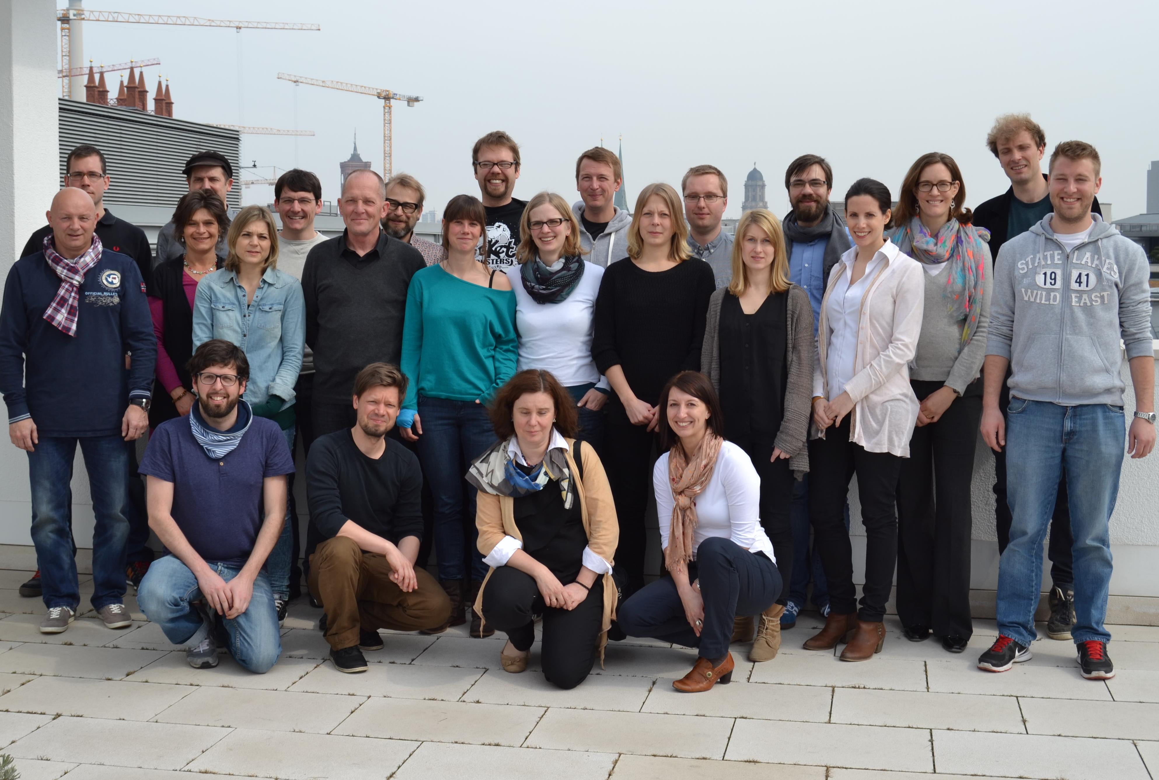 Eine Gruppe von etwa 30 Personen steht oder hockt lächelnd für ein Gruppenfoto zusammen. Im Hintergrund ist eine Stadtlandschaft mit modernen Gebäuden und Baukränen zu sehen. Die Personen sind unterschiedlich gekleidet, was auf eine vielfältige Gruppe hindeutet.
