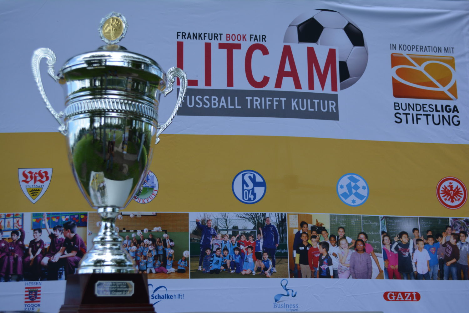 DFB-Pokal steht im Mittelpunkt eines Werbebanners mit dem Motto "Fußball trifft Kultur" auf der Frankfurter Buchmesse. Darunter sind Fotos von Kindern und Jugendlichen aus verschiedenen Fußballvereinen zu sehen, die an Projekten der DFL Stiftung teilnehmen.