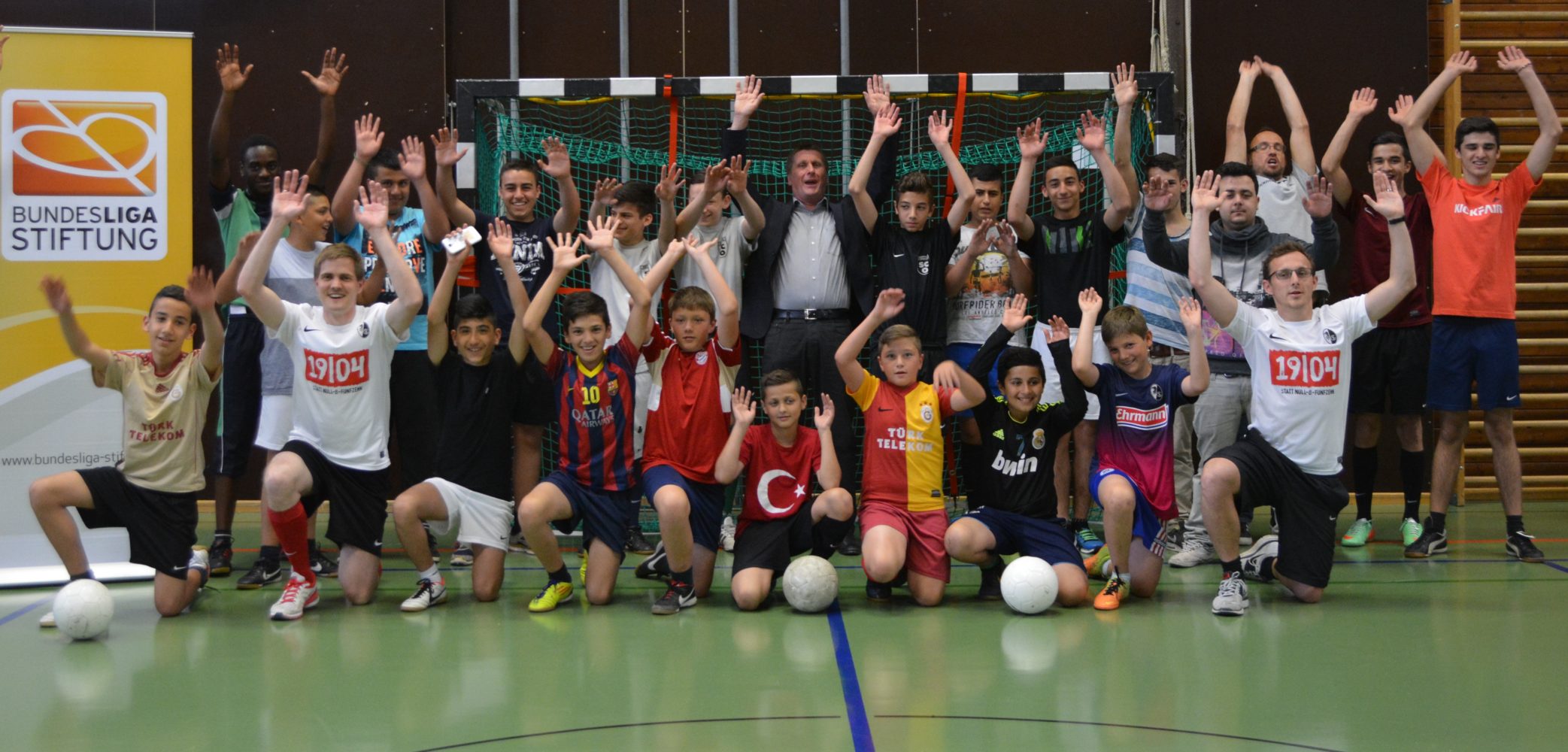 Eine Gruppe von Kindern und Jugendlichen steht in einer Sporthalle vor einem Handballtor und jubelt mit erhobenen Armen. Viele der Kinder tragen Trikots verschiedener Fußballvereine, darunter auch das Trikot der türkischen Nationalmannschaft. Im Hintergrund ist das Logo der DFL Stiftung zu sehen.