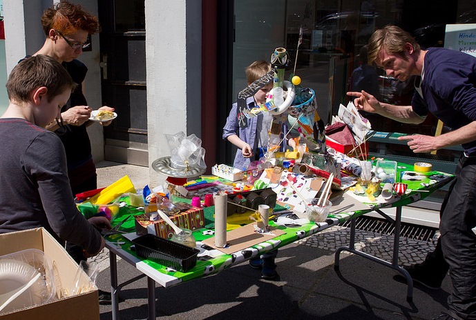 Kinder und Erwachsene basteln gemeinsam an einem bunt dekorierten Tisch im Freien. Auf dem Tisch befinden sich viele verschiedene Bastelmaterialien und kleine, selbstgebastelte Objekte.