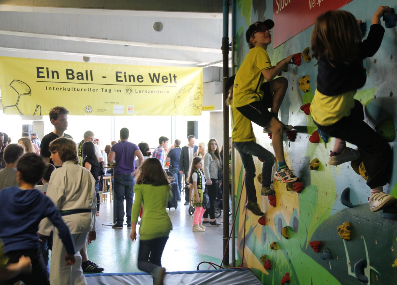 Eine Gruppe von Kindern und Jugendlichen nimmt an einem interkulturellen Tag im Lernzentrum des BVB teil. Ein Junge klettert an einer Kletterwand, während andere Kinder und Jugendliche im Hintergrund stehen und zuhören. Im Hintergrund ist ein Banner mit der Aufschrift „Ein Ball – Eine Welt“ zu sehen.