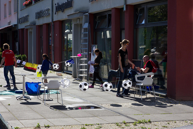 Fußball-Installation im öffentlichen Raum: Auf einem Gehweg stehen Stühle und um sie herum sind mehrere Fußballbälle verteilt. Im Hintergrund sind Menschen zu sehen, die an der Installation vorbeigehen.