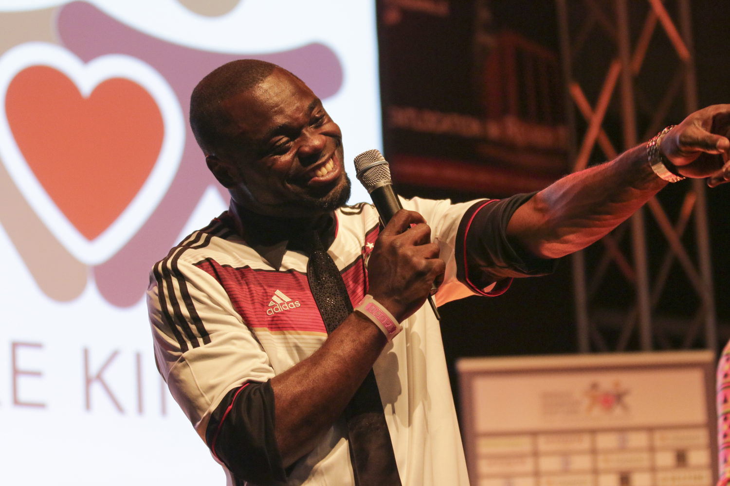 Ein lächelnder Mann mit dunkler Haut hält ein Mikrofon in der Hand und deutet mit ausgestrecktem Arm nach rechts. Er trägt ein weißes T-Shirt mit roten und schwarzen Adidas-Streifen. Im Hintergrund ist ein rotes Herz und die Buchstaben "E KI" zu sehen.