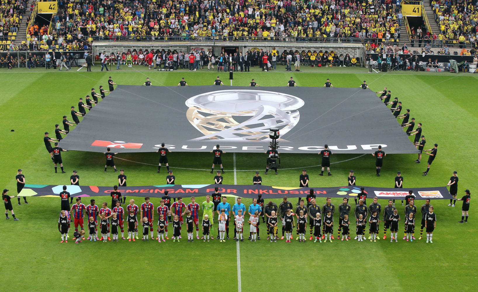 Fußballspieler beider Mannschaften, Borussia Dortmund und FC Bayern München, stehen im Kreis auf dem Rasen und halten ein großes Banner mit der Aufschrift "Gemeinsam für Inklusion" und dem Logo der Bundesligastiftung und Aktion Mensch. Im Hintergrund sind die Zuschauer auf den Tribünen zu sehen.
