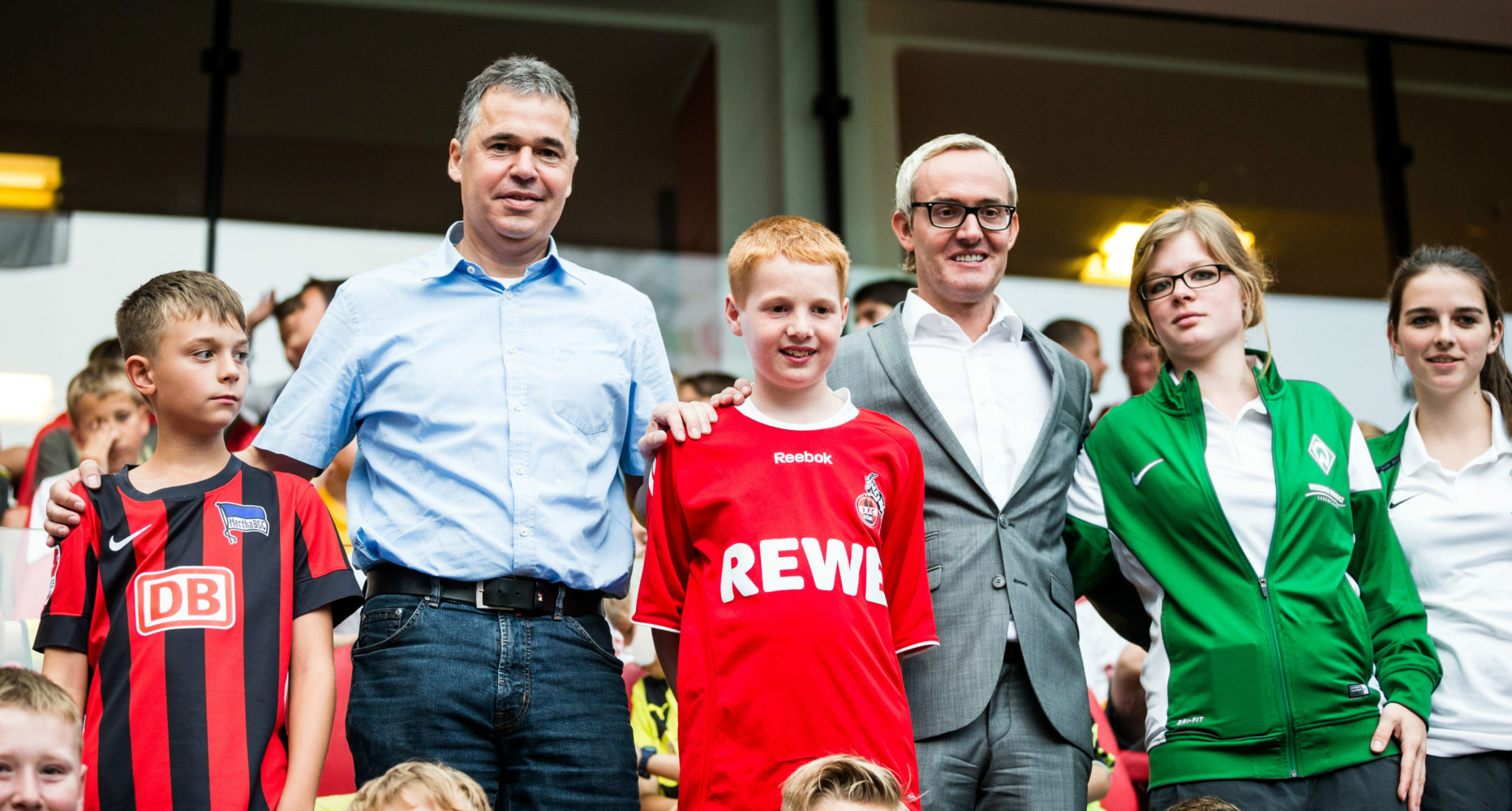 Zwei Jungen im Fußballtrikot stehen neben zwei Männern und einer Frau im Anzug auf einer Tribüne. Im Hintergrund sind weitere Personen und die Zuschauerränge zu sehen.