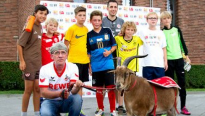 Eine Gruppe von Kindern und Erwachsenen posiert mit Hennes VIII, dem Ziegenbock des 1. FC Köln, vor einer Werbebande. Ein Mann in Vereinskleidung kniet vor dem Tier.