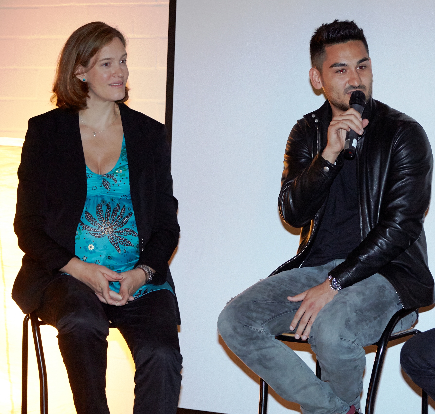 Zwei Personen sitzen auf Stühlen und führen ein Gespräch. Links eine Frau mit dunklem Blazer und blauem Oberteil, rechts ein Mann mit Lederjacke und Mikrofon in der Hand. Im Hintergrund eine weiße Wand.