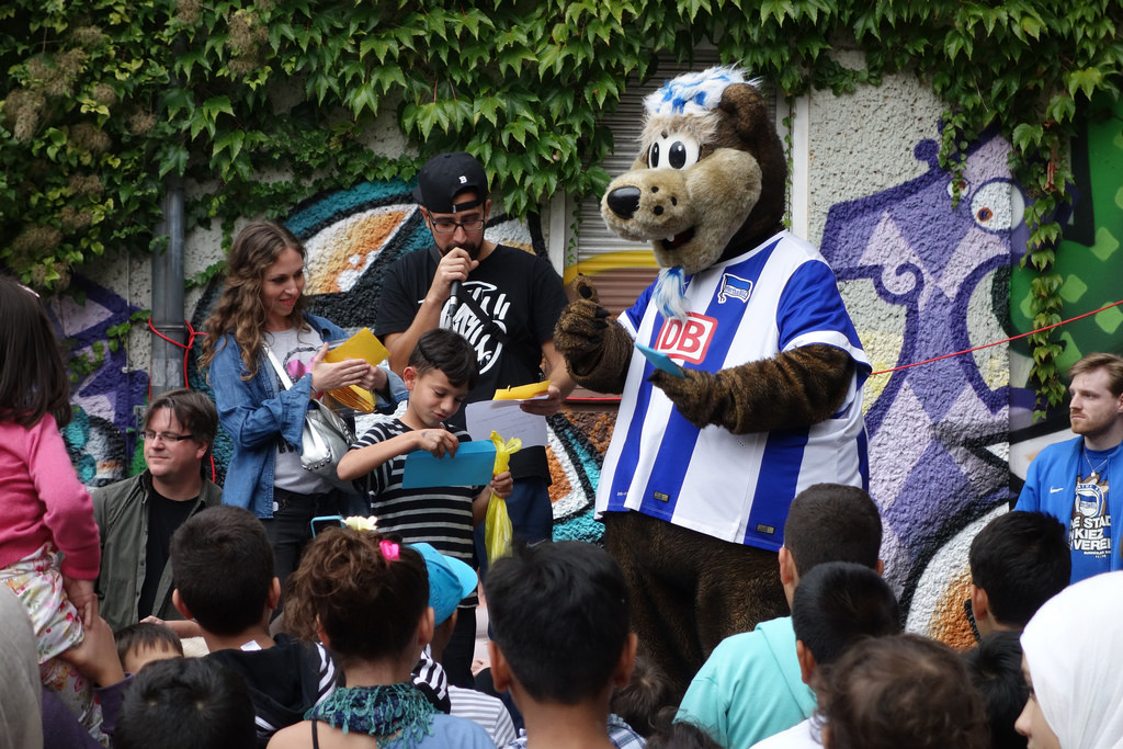 Eine Gruppe von Kindern und Erwachsenen umringt eine große Bären-Maskotte in Fußballkleidung. Die Bären-Maskotte nimmt ein Autogramm von einem jungen Fan entgegen, während andere Kinder und Erwachsene neugierig zusehen. Im Hintergrund ist eine Wand mit einem bunten Graffiti zu sehen.