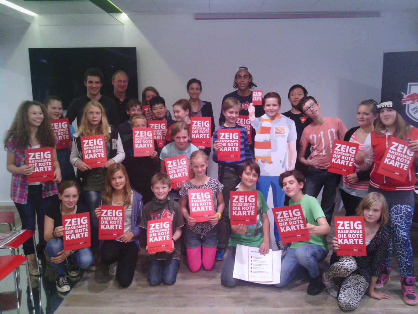 Eine große Gruppe von Jugendlichen und Erwachsenen hält rote Karten mit dem Aufdruck "ZEIG ROTE KARTE - RASSISMUS" in die Kamera. Sie stehen vor einem dunkelgrauen Hintergrund.