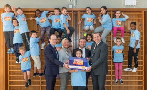 Eine Gruppe von Kindern in blauen T-Shirts und sportlicher Kleidung posiert gemeinsam mit drei Erwachsenen in einem Turnsaal. Ein Erwachsener übergibt den Kindern eine blaue Box.