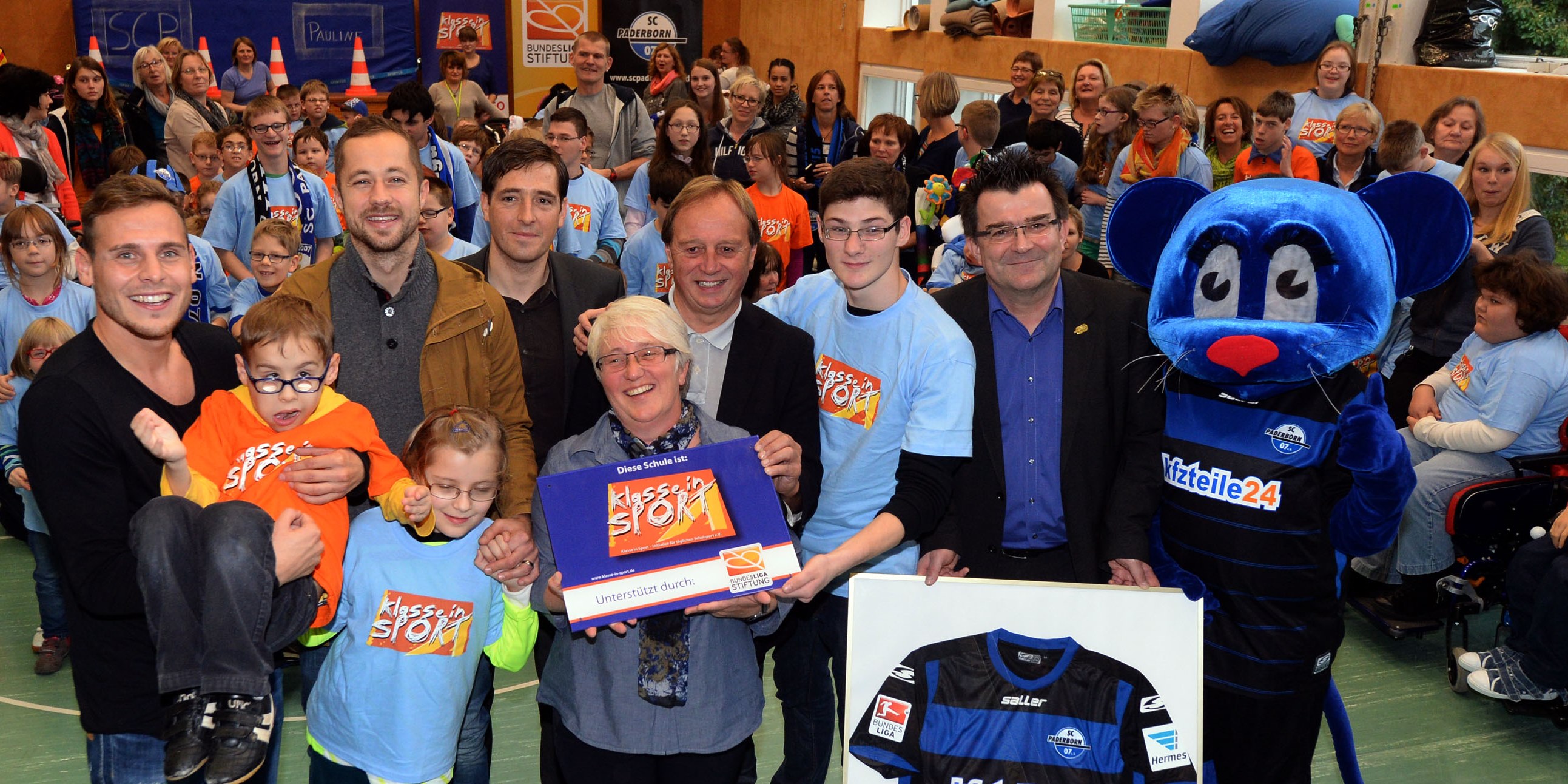 Gruppenfoto in einer Turnhalle: Schüler, Lehrer und Vertreter der DFL Stiftung und des SC Paderborn stehen zusammen. In der Mitte hält eine Schülerin eine Urkunde mit dem Aufdruck "Klasse in Sport". Ein Maskottchen des SC Paderborn steht am rechten Bildrand und hält ein Trikot.