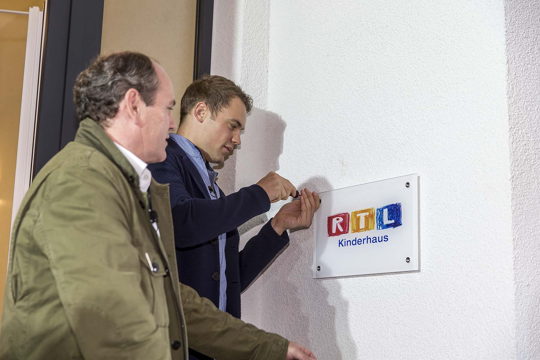 Ein Mann im blauen Blazer und ein anderer in einer Jacke stehen vor einem Gebäude. An der Wand hängt ein Schild mit der Aufschrift "Kinderhaus" in bunten Buchstaben.
