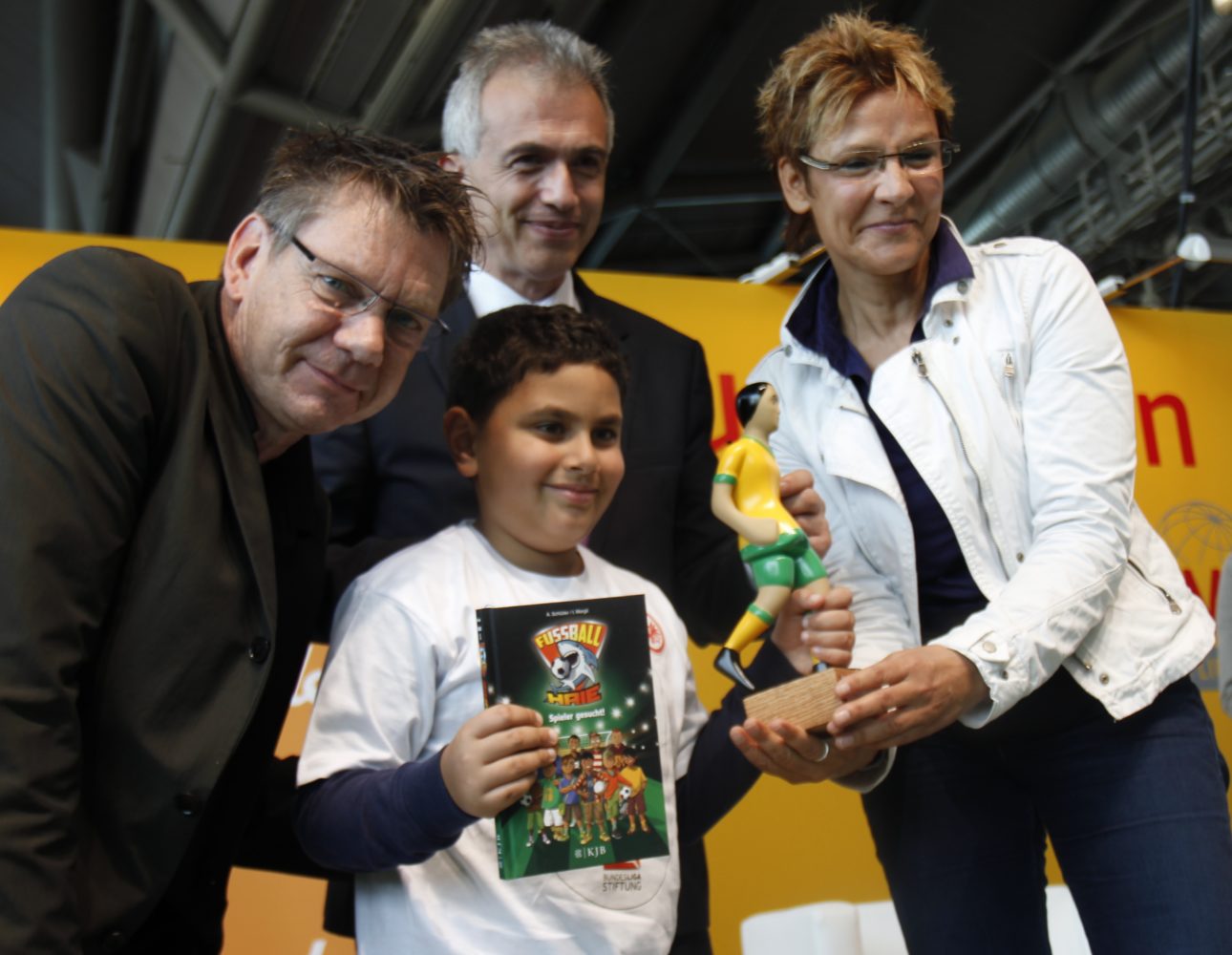 Ein Junge im weißen T-Shirt hält ein Buch mit dem Titel "Fußball Helden" in den Händen. Links steht ein Mann im schwarzen Hemd und schaut dem Jungen über die Schulter zu. Rechts steht eine Frau in einem weißen Jackett und überreicht dem Jungen eine kleine Figur eines Fußballspielers. Im Hintergrund ist ein Banner mit roten Buchstaben zu sehen.