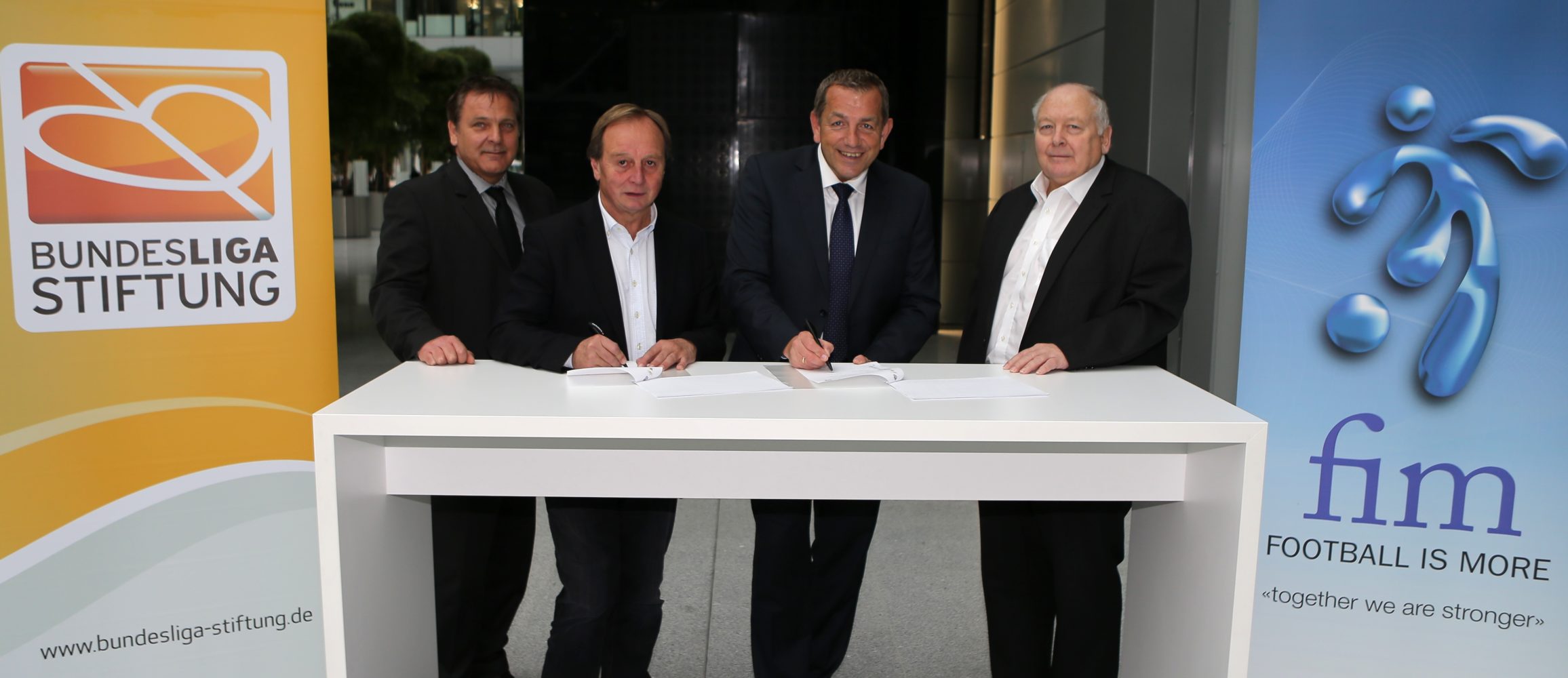 Vier Männer in Anzug und Krawatte sitzen an einem weißen Tisch und unterzeichnen ein Dokument. Im Hintergrund sind das Logo der Bundesliga Stiftung und das Logo von "FIM - FOOTBALL IS MORE - together we are stronger" zu sehen.