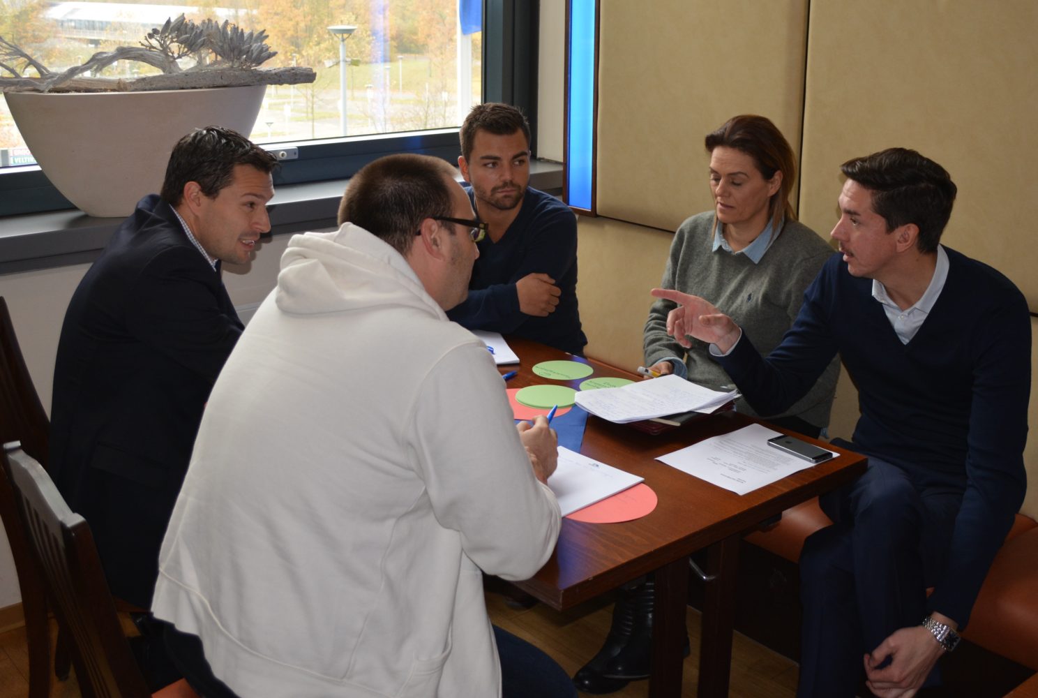 Fünf Personen sitzen an einem Tisch in einem hellen Raum und diskutieren. Eine Person im Hoodie sitzt dem Rest gegenüber und gestikuliert lebhaft. Die anderen Teilnehmer hören aufmerksam zu und tauschen Blicke aus. Auf dem Tisch liegen Papier und farbige Karten.