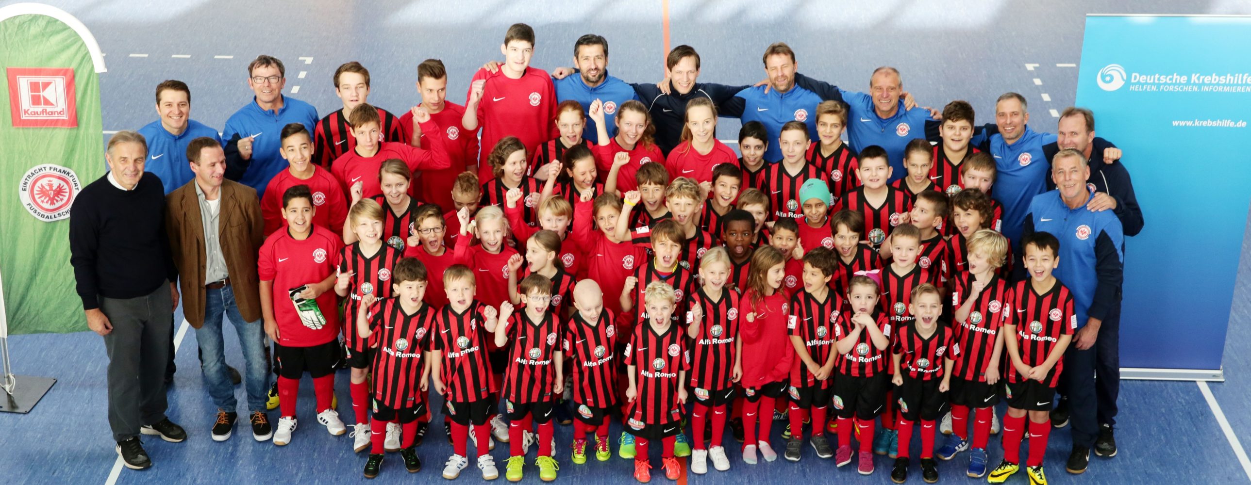 Gruppenfoto von Kindern und Jugendlichen in roten Trikots, umgeben von Betreuern und ehrenamtlichen Helfern in blauen Poloshirts. Im Hintergrund eine grüne Wand und Banner von Sponsoren.