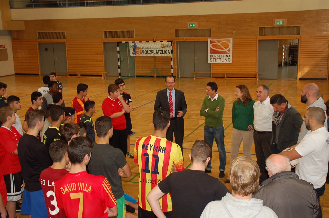 Eine Gruppe von Männern und Jugendlichen in Sportkleidung sitzt im Kreis auf dem Boden einer Sporthalle. Ein Mann in Anzug und Krawatte steht im Zentrum und scheint eine Ansprache zu halten. Im Hintergrund ist ein Banner mit der Aufschrift "BOLZPFLIGALIGA" und das Logo der DFL Stiftung zu sehen.
