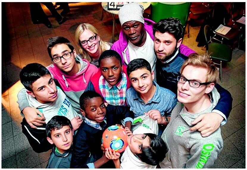 Eine Gruppe junger Menschen, darunter Kinder und Jugendliche mit unterschiedlichem Hintergrund, posiert gemeinsam und lächelt in die Kamera. Im Mittelpunkt hält ein Kind einen Fußball. Sie scheinen eine Gemeinschaft zu bilden und Freude zu empfinden.