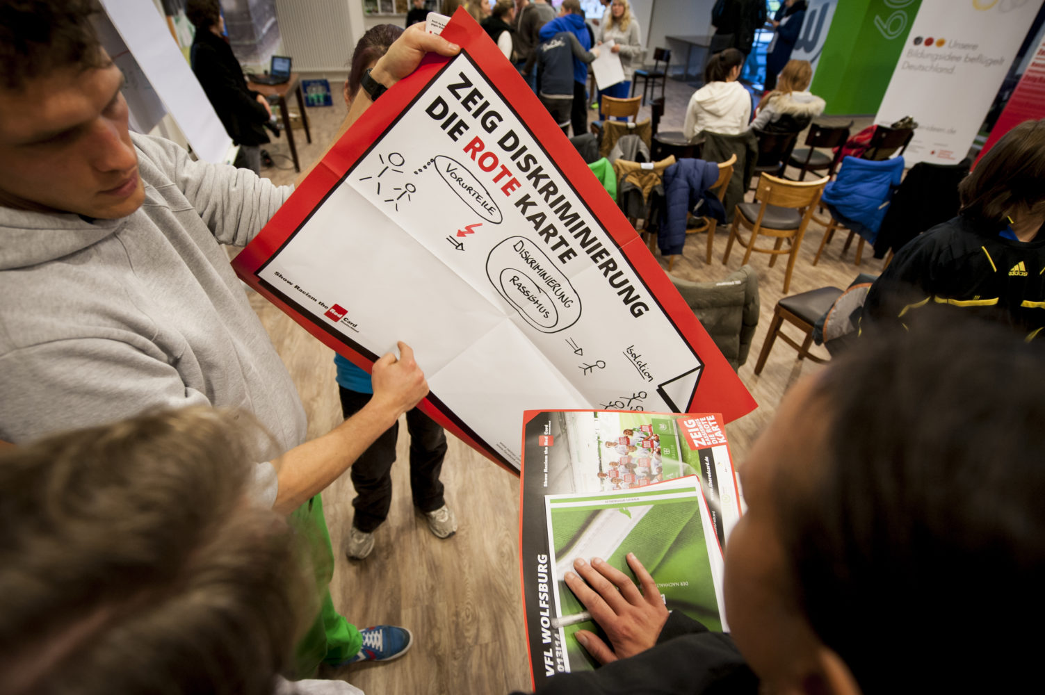 Ein Mann hält eine große rote Karte mit der Aufschrift "Zeig Diskriminierung die Rote Karte" in die Höhe. Im Hintergrund sind weitere Personen und ein Raum mit Tischen zu sehen. Die Szene zeigt die Preisverleihung des Projekts "Rote Karte für Rassismus", das im bundesweiten Wettbewerb "Ideen für die Bildungsrepublik" ausgezeichnet wurde.