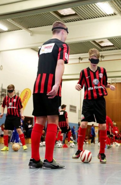 Fußballcamp: Zwei Jugendliche mit Augenbinde versuchen, einen Fußball zu spielen. Im Hintergrund weitere Kinder und Jugendliche, die ebenfalls an der Übung teilnehmen.