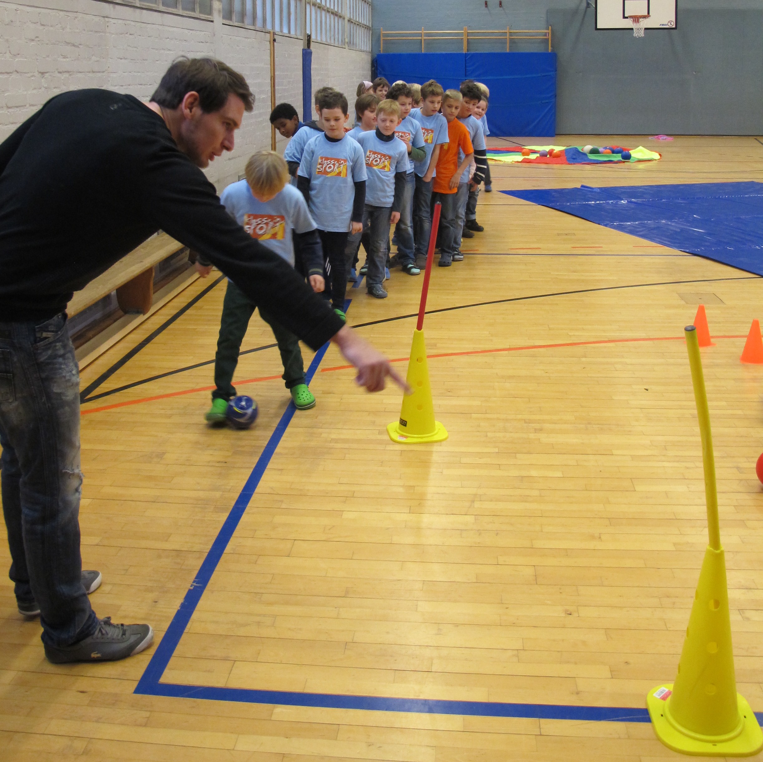 Ein Mann steht in einer Turnhalle und erklärt einer Reihe von Kindern, die in blauen T-Shirts gekleidet sind, eine Übung mit Hütchen. Die Kinder stehen auf einer blauen Linie auf dem Hallenboden.