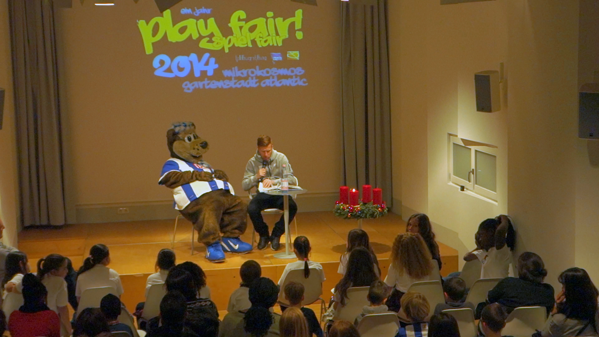 Ein Mann liest vor einer Gruppe von Kindern vor, während die Maskottchenfigur des FC Bayern München neben ihm sitzt. Im Hintergrund eine Bühne mit der Aufschrift "Play fair!" und "2014".