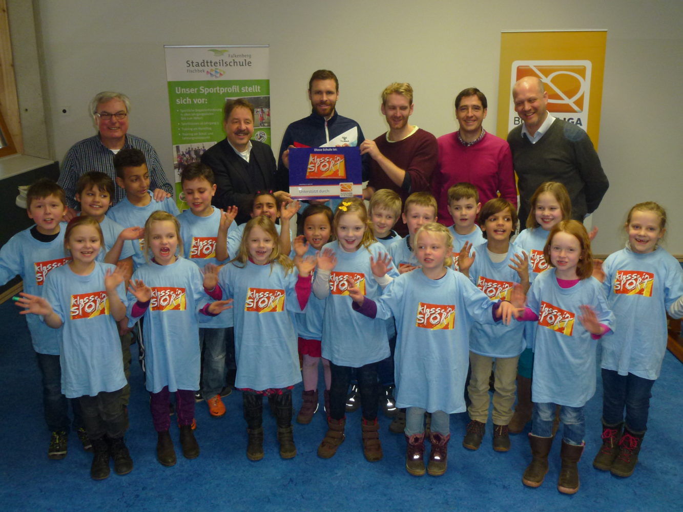 Eine Gruppe von Kindern in blauen T-Shirts mit dem Aufdruck "Klasse Sport" steht lächelnd in einer Turnhalle. Hinter ihnen stehen Erwachsene, darunter auch der Fußballspieler Marc Stendera vom FC St. Pauli, der eine Urkunde hält. Im Hintergrund ist ein Banner der Stadttheilschule zu sehen.