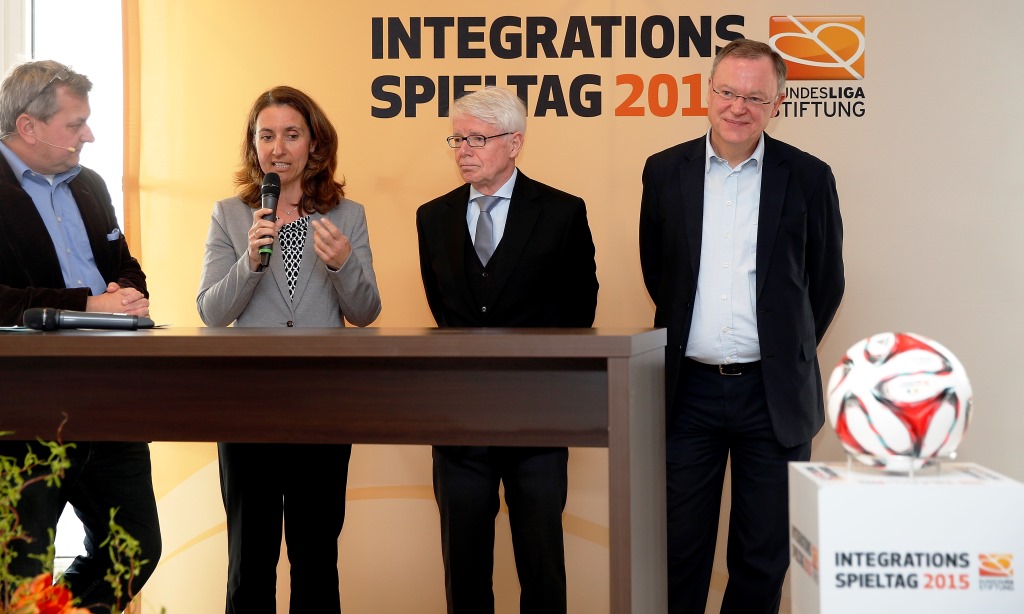 Eine Frau hält ein Mikrofon und spricht vor einem Podium. Neben ihr stehen zwei Männer in Anzügen. Im Hintergrund ist ein Banner mit der Aufschrift "INTEGRATIONS SPIELTAG 2015" und dem Logo der Bundesliga-Stiftung zu sehen. Rechts steht ein Fußball auf einem Podest.