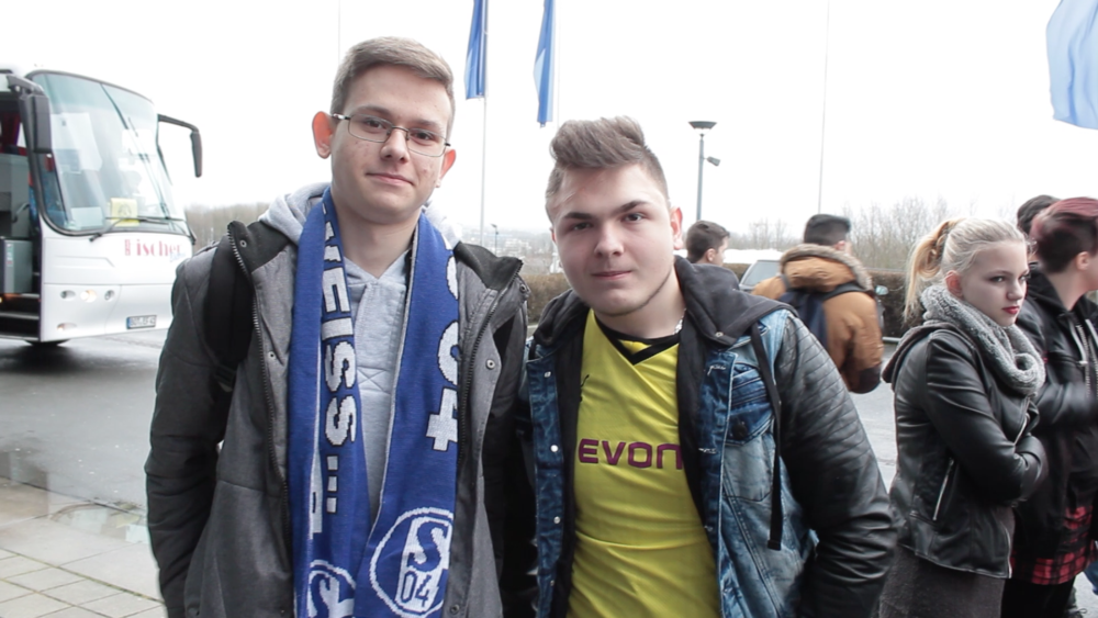 Zwei junge Männer stehen vor einer Menschenmenge und lächeln in die Kamera. Der Mann links trägt einen blauen Schal mit dem Logo "S04" und eine schwarze Jacke. Der Mann rechts trägt ein gelbes Trikot mit dem Aufdruck "Evonik" und eine Jeansjacke. Im Hintergrund ist ein Reisebus und weitere Personen erkennbar.
